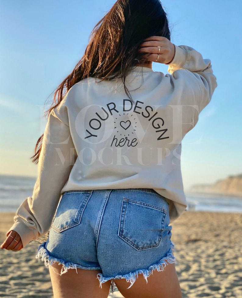 以下が含まれることがあります： 女性は、背中に「YOUR DESIGN here」と書かれた薄いベージュのトレーナーを着ています。彼女は青いデニムのショートパンツを履いており、砂浜に立っています。