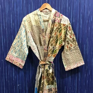 May include: A patchwork robe with a cream-colored base and colorful, geometric patterns. The robe is hanging on a wooden hanger against a blue background. The robe has long sleeves and a tie belt.
