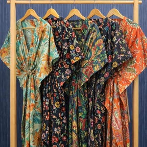 Puede incluir: Cinco batas con estampado floral colgadas en un perchero de madera. Las batas presentan varios patrones con colores como naranja, azul, verde y negro. Cada bata tiene cuello en V y lazo en la cintura.