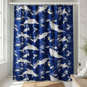 Puede incluir: Una cortina de ducha azul marino con un patrón repetido de delfines blancos y peces amarillos. Burbujas blancas están dispersas por todo el diseño. La cortina cuelga en un baño, con una ventana y una alfombra de baño de madera visibles.