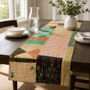 May include: A rectangular table runner featuring a patchwork design of various fabric squares in shades of orange, teal, green, and cream. The runner is decorated with intricate white kantha stitching and floral patterns, laid flat across a dark wooden table.