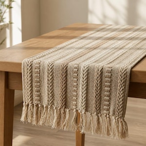 May include: A rectangular table runner featuring a woven texture with alternating vertical stripes in shades of beige, tan, and cream. The fabric displays a raised cable-knit pattern and is finished with long, knotted fringe tassels on both ends.