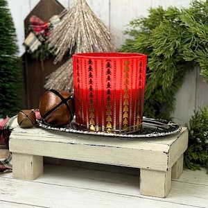 Puede incluir: Un portavelas de cristal rojo con diseños de árboles de Navidad dorados. El portavelas se encuentra sobre una bandeja plateada con dos campanas marrones. La bandeja está sobre un pequeño soporte de madera blanco. Hay vegetación y una pieza decorativa de madera en el fondo.