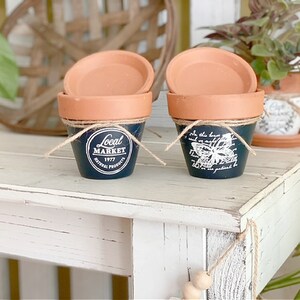 Könnte beinhalten: Zwei kleine Terrakotta-Blumentöpfe mit passenden Untertassen. Die Töpfe sind marineblau lackiert und mit einem weißen Schmetterlingsdesign und dem Text "Local Market 1977 Natural Products" verziert. Jeder Topf ist mit Bindfaden gebunden.