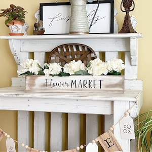 Peut inclure: Une étagère en bois blanc vieilli présente une enseigne "Flower Market" avec des fleurs blanches. Décor de style ferme avec un panier tressé, des cadres et un pichet en métal. Une guirlande avec des étiquettes et des perles en bois est suspendue en dessous.