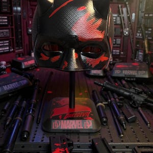 May include: A black Daredevil mask with red accents and horns, displayed on a stand. The mask has a textured surface and is set against a background of various props. The stand features the Daredevil and Marvel logos.