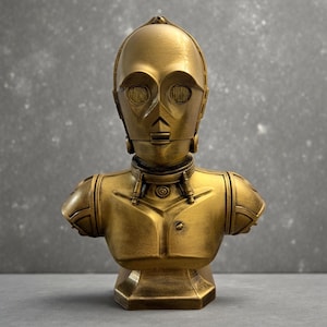 May include: A gold-colored bust of the droid C-3PO, featuring a metallic finish, a rounded head with circular eye sockets, a neck collar with visible mechanical details, and a chest plate with a small circular indentation on the left side.