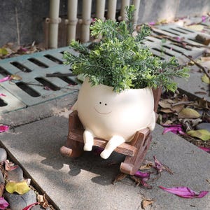 May include: A cream-colored, round planter shaped like a character with a simple smiling face, seated in a small brown wooden-style rocking chair. The planter holds a bushy, vibrant green artificial plant with needle-like foliage.