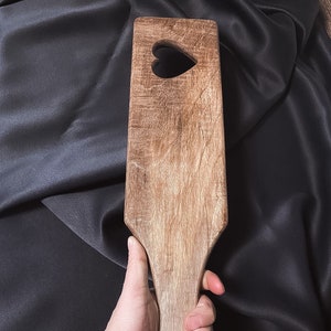 Puede incluir: Una paleta de madera con un recorte en forma de corazón. La paleta tiene un mango con un anillo en el extremo.