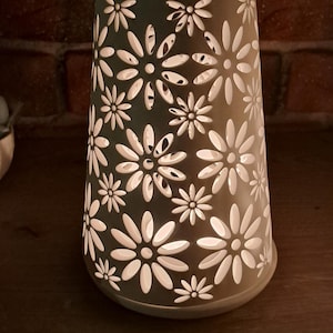 Könnte beinhalten: Eine konische Lampe mit einer matten grauen Außenseite, die ein sich wiederholendes Ausschnittmuster aus weißen Gänseblümchen aufweist. Die Lampe strahlt ein sanftes, warmes Licht von innen aus und beleuchtet die Blumenformen.