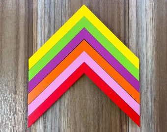May include: A colorful arrow shape made of six layers of wood painted in different colors: yellow, green, purple, orange, pink, and red.