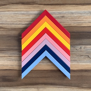 May include: A colourful chevron-shaped wall decor made of multiple layers of painted wood. The colours are red, orange, yellow, pink, purple, blue, and navy blue.