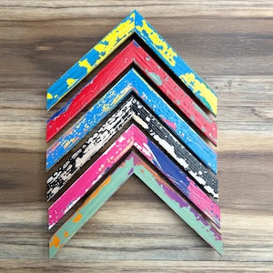 May include: A collection of colourful, distressed wooden picture frames arranged in a chevron pattern. The frames feature vibrant hues of blue, red, black, pink, and green with a weathered, textured finish, set against a wood grain background.