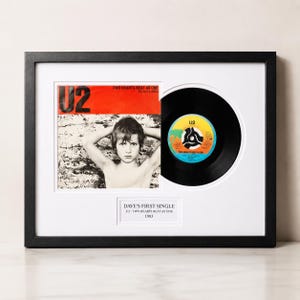 May include: A framed display featuring a U2 Two Hearts Beat As One vinyl record and its original sleeve. The sleeve shows a person with hands behind their head. A white plaque below reads DAVE'S FIRST SINGLE U2 - TWO HEARTS BEAT AS ONE 1983.