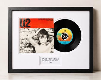 May include: A framed display featuring a U2 Two Hearts Beat As One vinyl record and its original sleeve. The sleeve shows a person with hands behind their head. A white plaque below reads DAVE'S FIRST SINGLE U2 - TWO HEARTS BEAT AS ONE 1983.