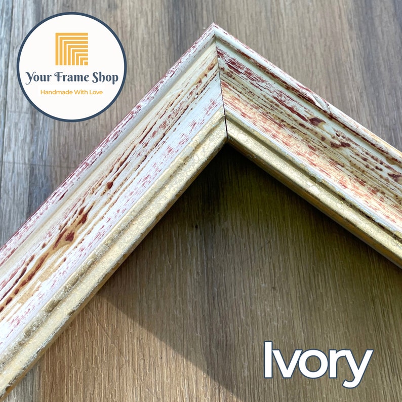 Puede incluir: Primer plano de una esquina de un marco de imagen blanco y dorado con un acabado desgastado y envejecido. El marco tiene un borde elevado y un sutil tono rojo. El texto "Your Frame Shop Handmade With Love" es visible en la esquina superior izquierda. La palabra "Ivory" es visible en la esquina inferior derecha.