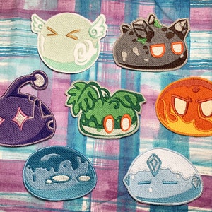 May include: Eight embroidered patches featuring different colored slime-like creatures with various facial expressions. The patches are on a purple and blue plaid fabric background.