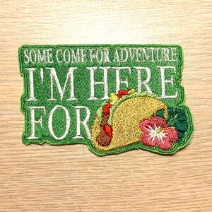Puede incluir: Parche bordado verde con el texto "Some come for adventure I'm here for" y un taco bordado amarillo y marrón con flores bordadas verdes y rojas.