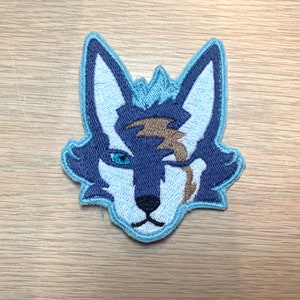 May include: Embroidered patch featuring a stylized wolf head. The wolf has blue and white fur with a brown lightning bolt-shaped scar over one eye. The patch is outlined in light blue and is likely for clothing or accessories.