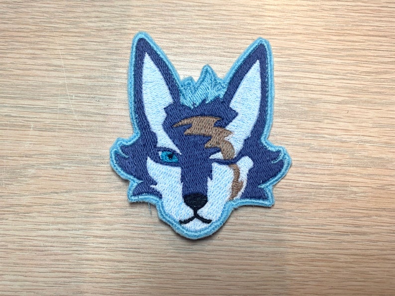May include: Embroidered patch featuring a stylized wolf head. The wolf has blue and white fur with a brown lightning bolt-shaped scar over one eye. The patch is outlined in light blue and is likely for clothing or accessories.
