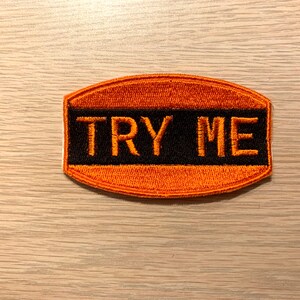 May include: Orange and black embroidered patch with the text "TRY ME" in white.
