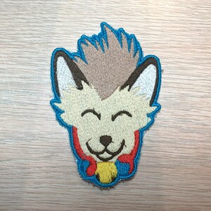 May include: Embroidered patch featuring a smiling cartoon fox with a light tan face, brown ears, and a blue outline. The fox has a light brown tuft of hair and a red and yellow collar. The patch is designed for apparel or accessories.