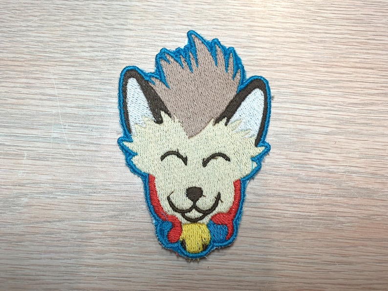May include: Embroidered patch featuring a smiling cartoon fox with a light tan face, brown ears, and a blue outline. The fox has a light brown tuft of hair and a red and yellow collar. The patch is designed for apparel or accessories.