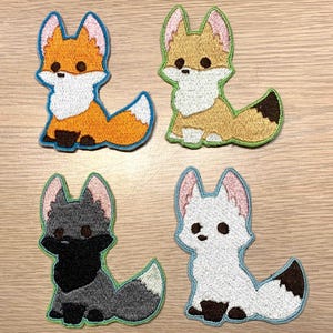May include: Four embroidered iron-on patches featuring different colored foxes. The foxes are all in a cartoon style with big eyes and small mouths. The patches are all about 2 inches tall and 2 inches wide.