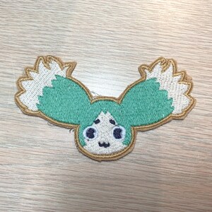 May include: Embroidered patch featuring a cartoon character with teal hair and white accents. The patch has a tan border and a smiling face with black eyes and a small mouth. The design is on a light wood grain surface.