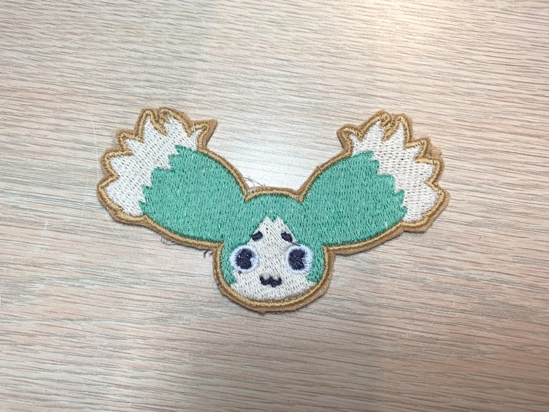 May include: Embroidered patch featuring a cartoon character with teal hair and white accents. The patch has a tan border and a smiling face with black eyes and a small mouth. The design is on a light wood grain surface.