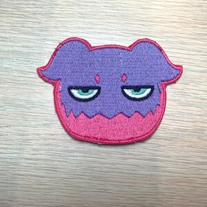 May include: Embroidered patch featuring a purple and pink cartoon character with a grumpy expression. The character has green eyes and a red outline. The patch is likely for clothing or accessories.