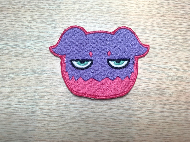 May include: Embroidered patch featuring a purple and pink cartoon character with a grumpy expression. The character has green eyes and a red outline. The patch is likely for clothing or accessories.