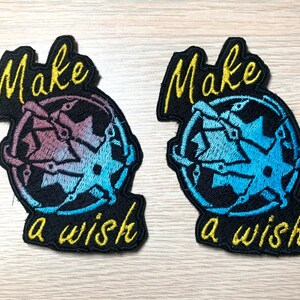 Può includere: Due toppe ricamate nere con il testo "Make a wish" in filo giallo. Le toppe presentano un design a stella blu e viola.