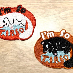 Op de afbeelding: Twee geborduurde strijkpatches met een witte kat en een zwarte kat die slapen op het woord "moe". De tekst "Ik ben zo" staat boven de kat.