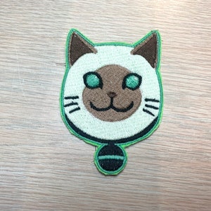 May include: Embroidered patch featuring a cartoon cat face with a white head, brown ears and nose, and green eyes. The cat has a green outline and a small black and green pendant. The patch is designed for sewing or ironing onto fabric.