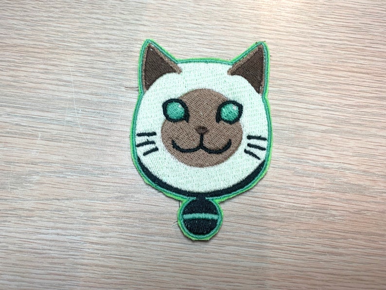 May include: Embroidered patch featuring a cartoon cat face with a white head, brown ears and nose, and green eyes. The cat has a green outline and a small black and green pendant. The patch is designed for sewing or ironing onto fabric.