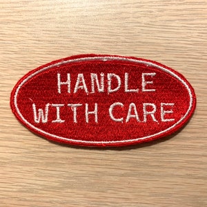 May include: Red oval-shaped patch with white embroidered text that reads "HANDLE WITH CARE".