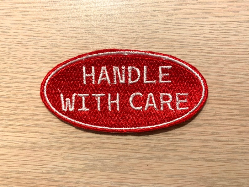 May include: Red oval-shaped patch with white embroidered text that reads "HANDLE WITH CARE".