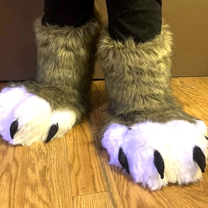 May include: Furry gray and white paw boots with black claws. The boots have a plush, furry texture and are designed to resemble animal paws. The boots are worn over black pants and are on a wooden floor.