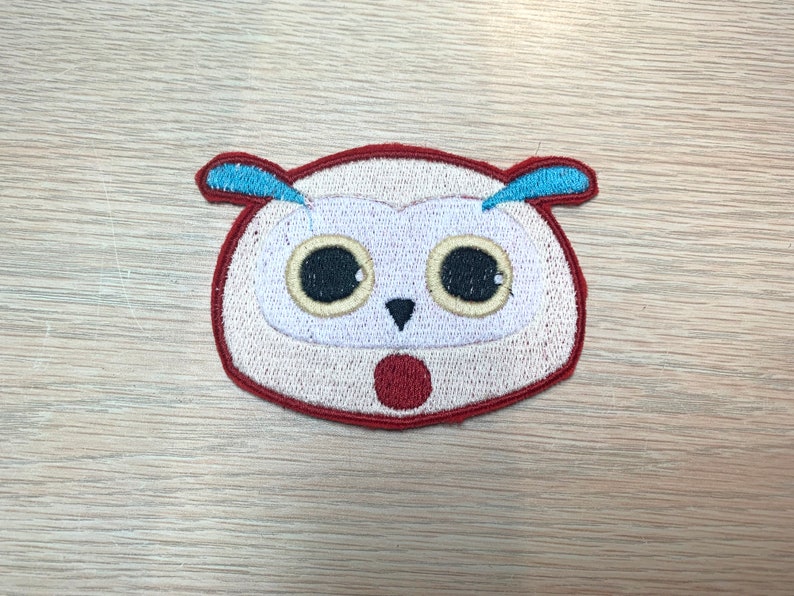 May include: Embroidered owl patch with a surprised expression. The owl has a white face, large black eyes, and a red open mouth. It features blue ear tufts and a red border. This patch is suitable for clothing or accessories.