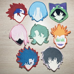 Puede incluir: Ocho parches bordados para planchar que representan diferentes personajes con varios colores y estilos de cabello. Los parches están sobre una superficie marrón claro.