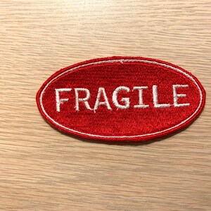 May include: Red oval-shaped fabric patch with white embroidered text that reads "FRAGILE".