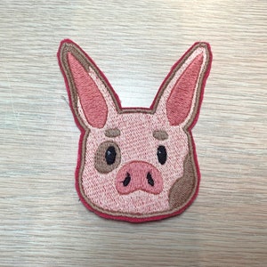 May include: Embroidered patch featuring a pig's head. The pig has pink skin, brown spots, and a red outline. The eyes are black, and the ears are long and pointed. This patch is suitable for jackets, bags, or other fabric items.