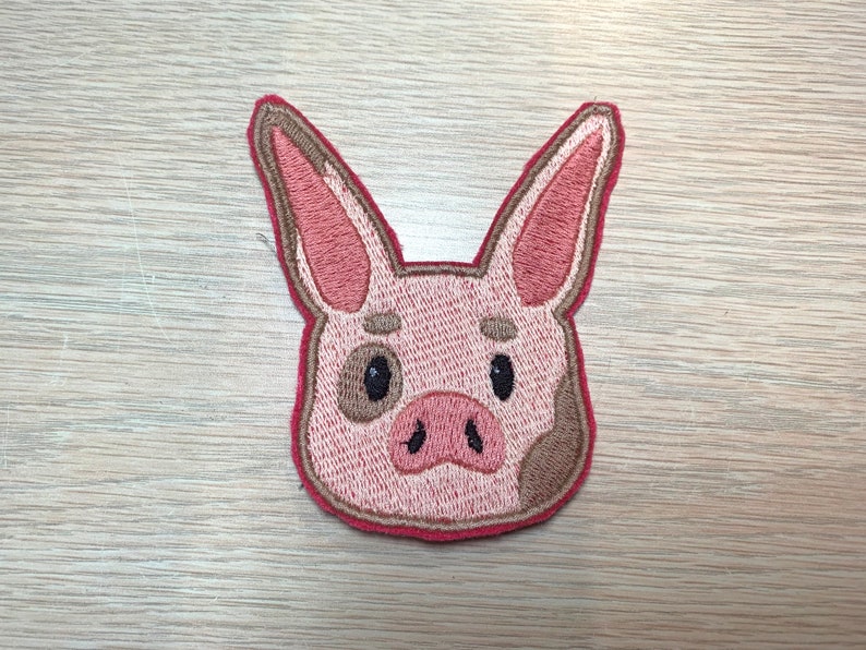 May include: Embroidered patch featuring a pig's head. The pig has pink skin, brown spots, and a red outline. The eyes are black, and the ears are long and pointed. This patch is suitable for jackets, bags, or other fabric items.