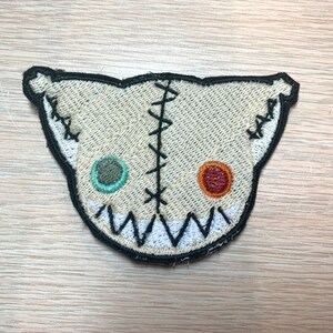 May include: A beige, stitched patch in the shape of a stylized cat face. The patch features a black outline, black stitches, and two different colored eyes. The mouth is a jagged white line.