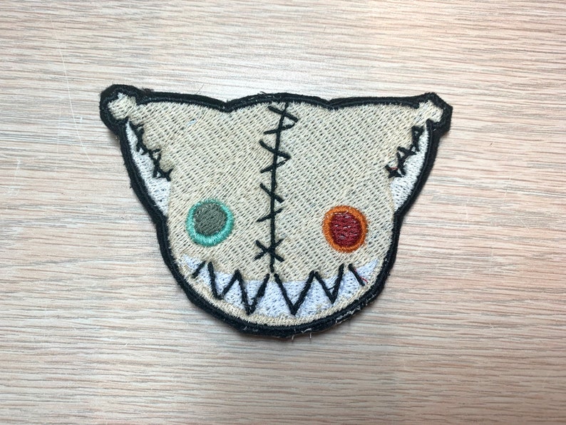 May include: A beige, stitched patch in the shape of a stylized cat face. The patch features a black outline, black stitches, and two different colored eyes. The mouth is a jagged white line.