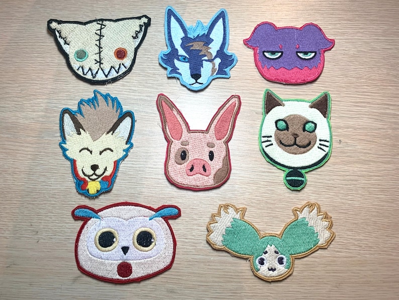 May include: A collection of nine embroidered patches featuring various cartoon animal faces. The patches include a stitched-together doll head, a wolf, a purple creature, a smiling wolf-like character, a pig, a cat, an owl, and a stylized owl.