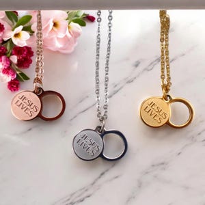 Op de afbeelding: Drie kettingen met de tekst "JESUS LIVES" op een ronde hanger. De kettingen zijn in roségoud, zilver en goudtinten. Elke hanger heeft een cirkelvormig ontwerp. De achtergrond heeft roze bloemen.