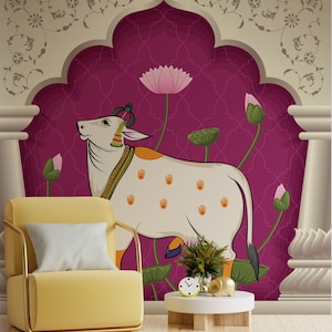 Op de afbeelding: Een decoratieve muurschildering met een gestileerde witte koe met oranje handafdrukken, versierd met sieraden en een pauwenveer. De koe staat tegen een magenta achtergrond met lotusbloemen en een bloemenboog. Een gele fauteuil en een bijzettafel staan op de voorgrond.