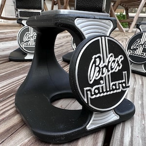 May include: Black and silver camera stands with the text "Bolex Paillard" in a circular design. The stands have a curved base and a platform for a camera. The stands are on a wooden surface.